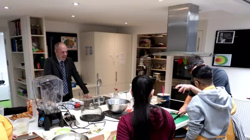 A photograph of Justin Tomlinson MP visiting the kitchen at the Centrepoint Dean Street centre, watching a cooking workshop with two young people.