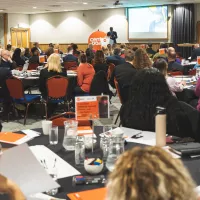 A large room with people at round tables listening to a Centrepoint speaker on the stage