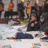 Two women sit in Sleep Out branded sleeping bags, one drinking a coffee