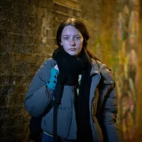 A young woman stands in a dimly lit alley with a brick wall covered in graffiti. She wears a thick grey puffer jacket, black scarf, and teal fingerless gloves, looking directly at the camera with a serious expression.