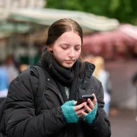 A young girl dressed in a thick black jacket and frayed blue gloves looks down at her phone. She carries a backpack and is alone, with a busy street behind her.