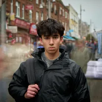 A young boy stares into the camera, holding a backpack over one shoulder and wearing a black jacket. He is still while there is lots of action on the street behind him, with people walking by. 