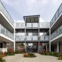 A modern three‑storey housing block with open walkways and metal railings surrounding a central courtyard filled with plants and benches.