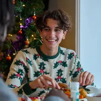 A young man sits at a table eating Christmas dinner, smiling at another person across from him. There is a Christmas tree behind him and he is wearing a Christmas jumper with holly on it. 