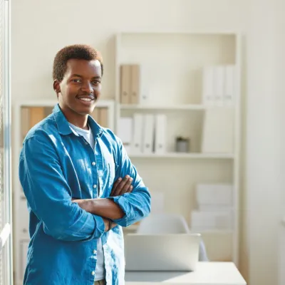 A young man standing confidently with arms crossed, wearing a blue button-up shirt over a white t-shirt. He is smiling at the camera in a bright office space, with shelves of organized files and a window seat in the background.