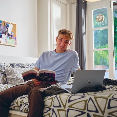 A young person sitting on a neatly made bed in a bright bedroom, holding an open book and smiling at the camera. A laptop rests on the bed, and a pinboard with notes and photos hangs on the wall behind them.