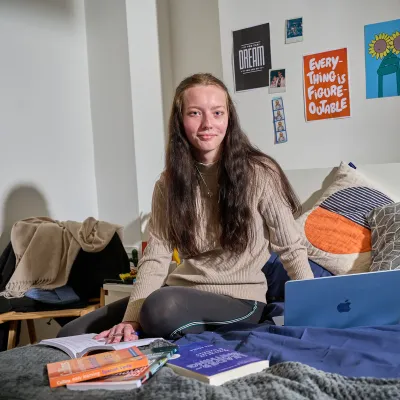 A young woman, NB, sits on a bed in her room, which is decorated with posters, coloured cushions, and books. She looks to the camera and has a smile on her face.