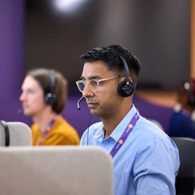 A Helpline Advice Worker wearing a headset and glasses sits at a computer in a call centre, focused on his screen, with other colleagues working at desks in the background.