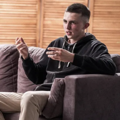 A person seated on a sofa in a living room speaks with another person seated opposite. The person on the sofa is gesturing with both hands while engaged in conversation. The second person is visible in the foreground, out of focus. Soft indoor lighting and wooden paneling create a warm, informal setting.  Provide your feedback on BizChat