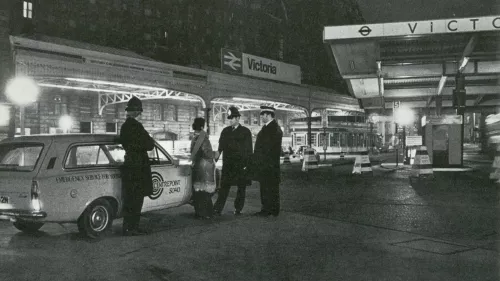 Black and white photograph showing three police officers speaking with a Centrepoint worker near a car with Centrepoint branding on it outside Victoria railway station in London