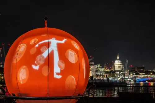 The snow globe art installation lights up in orange at night, showing a cartoon-style young person struggling with the pressures of homelessness. St Pauls Cathedral and the Thames are in the background