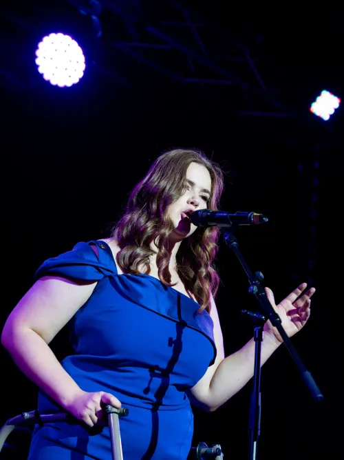 A young person with long hair, wearing a blue dress sings on stage