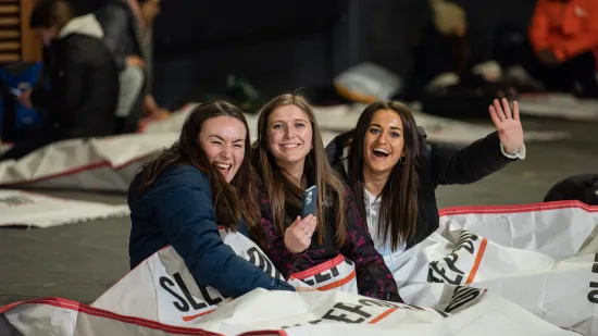 Three people sit on the ground inside a giant sleeping back waving at the camera