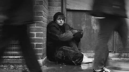 A black and white photo of Leo, sitting huddled on the floor in a doorway as people walk past quickly.