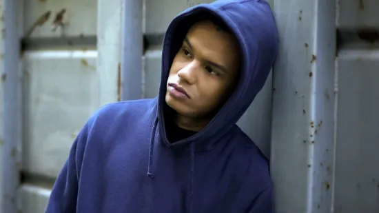 Young person in a blue hoodie leans against a fence