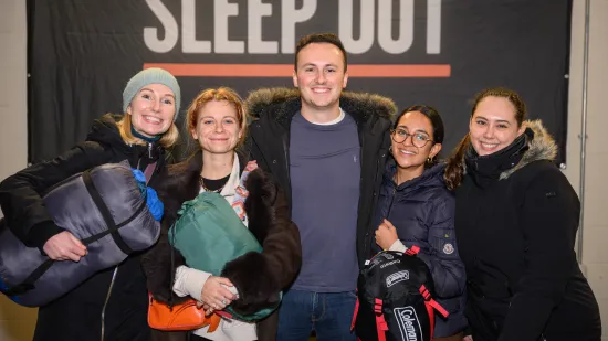 Five sleep out participants smile at the camera in front of a Sleep Out banner holding sleeping bags