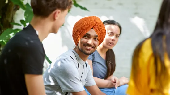 Young man is sat outside with young people and is smiling at another young person in the group.. 