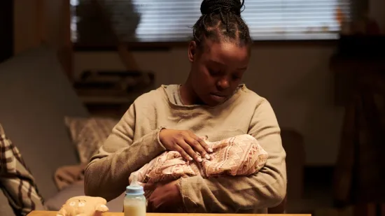 Young mother lovingly holding and caring for her newborn baby at home, with a baby bottle and stuffed toy nearby