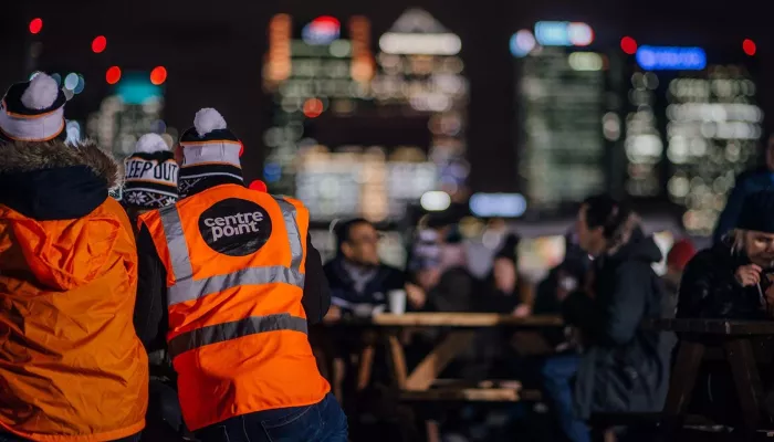 Two people stood with their backs to the camera wearing high vis jackets and bobble hats with London's skyline in the background.