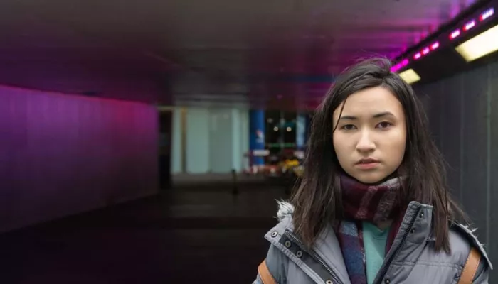 Homeless young person standing underneath an underpass, wearing a scarf and looking to the camera.