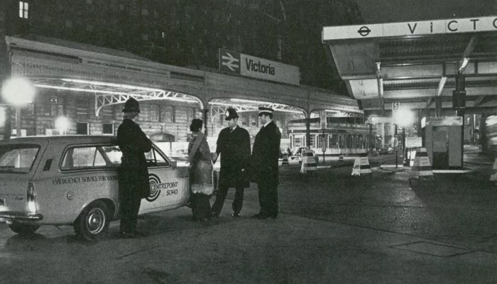 Black and white photograph showing three police officers speaking with a Centrepoint worker near a car with Centrepoint branding on it outside Victoria railway station in London