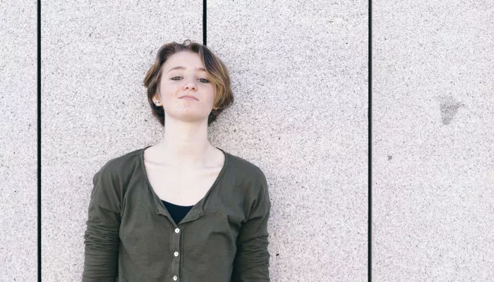 Young person with short hair leans against a grey wall 