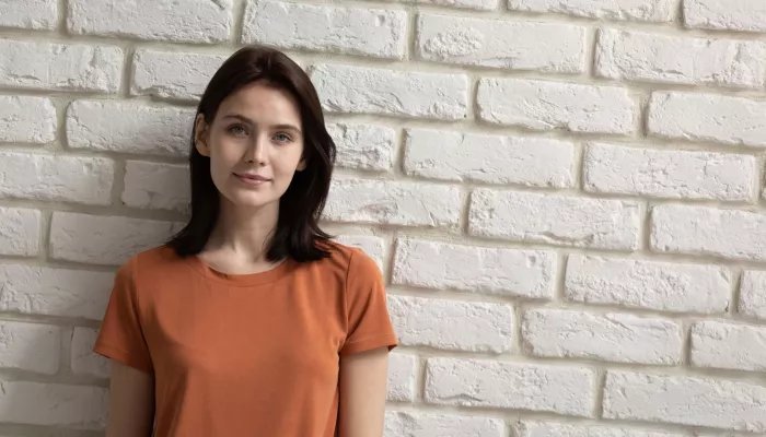 Young person with long dark hair in an orange t-shirt against a white brick wall