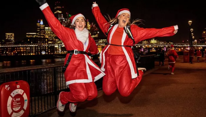 Photograph of two people by The Thames river jumping in Santa outfits