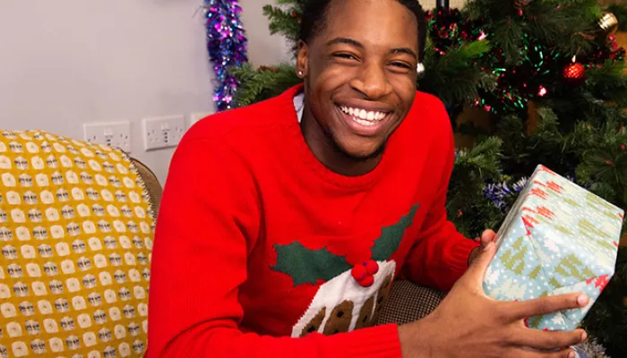 Young person smiling and holding a gift in front of a Christmas tree