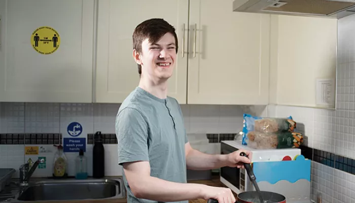 A young person smiles in a kitchen, cooking food in a red saucepan on a stove