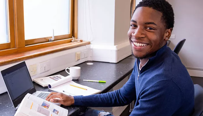 A young person sits at a desk with a laptop and notebooks, studying for their lessons
