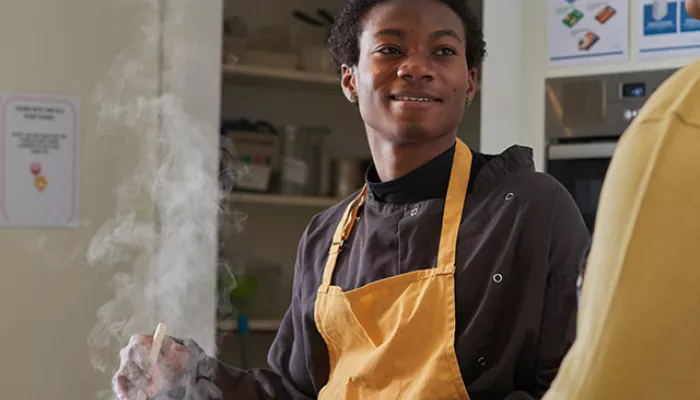 A young person stands in an apron while cooking food in a saucepan on a stove