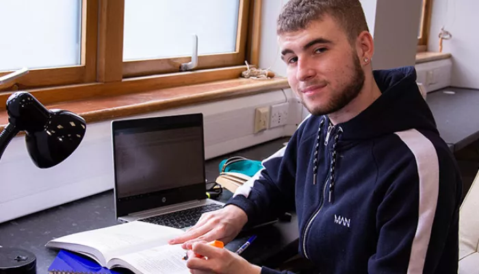 A young person sits at a desk with a laptop and notebooks, studying for their lessons
