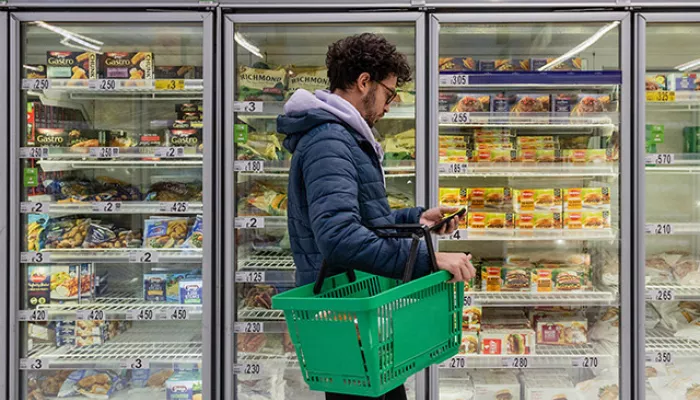 A young person holds a green shopping basket and browses a supermarket fridge