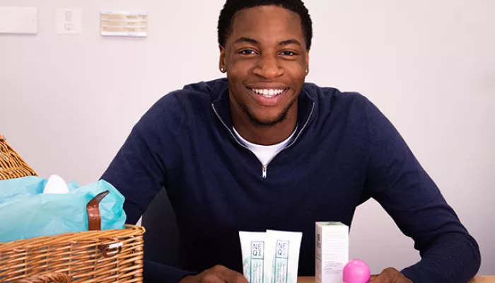 A young person warmly smiles at the camera, holding a variety of toiletries including deodorant and toothpaste