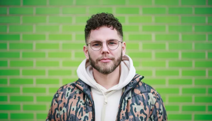 A portrait of Radio 1’s Dean McCullough directly facing the camera in front of a green brick wall