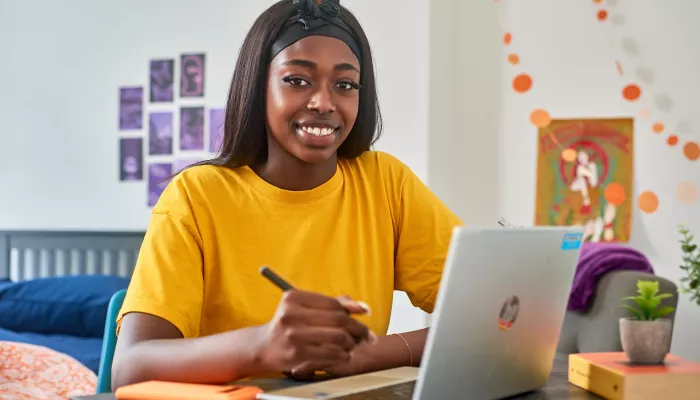 A young person sits at a desk with a laptop, pen and notepad in their own room