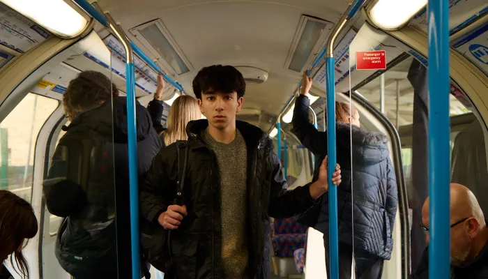 A young homeless person stands looking scared in a busy tube train carriage holding a bag of their belongings