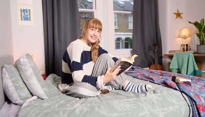 A young woman sits on a bed, reading and smiling.