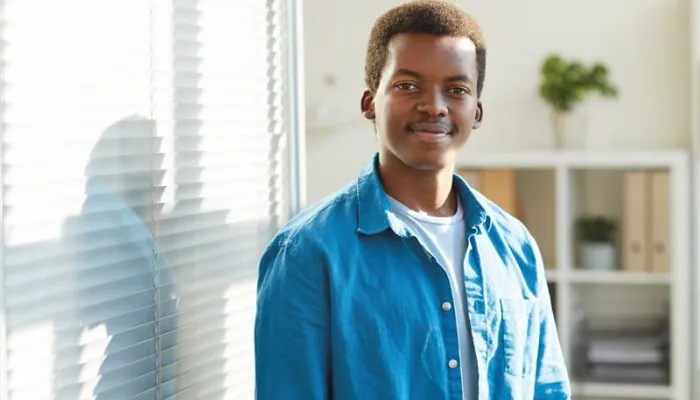 A young man standing next to a window with blinds, wearing a blue button-up shirt over a white t-shirt. He is smiling softly and looking at the camera, with a bookshelf and a potted plant in the background.