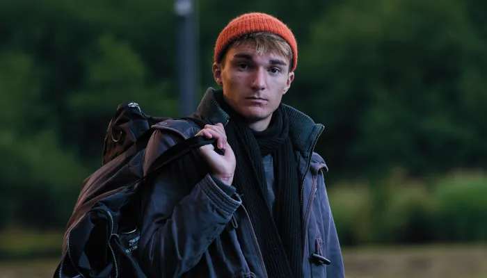A young person standing outdoors on a patch of grass, carrying a duffel bag over one shoulder and holding a rolled-up sleeping bag. They’re wearing a grey parka, green trousers, a scarf, and an orange beanie, with trees in the background.