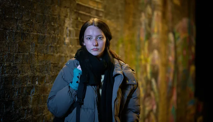 A young woman stands in a dimly lit alley with a brick wall covered in graffiti. She wears a thick grey puffer jacket, black scarf, and teal fingerless gloves, looking directly at the camera with a serious expression.