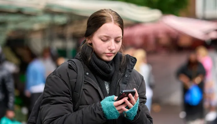 A young girl dressed in a thick black jacket and frayed blue gloves looks down at her phone. She carries a backpack and is alone, with a busy street behind her.
