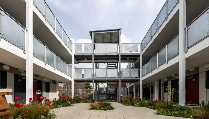 A modern three‑storey housing block with open walkways and metal railings surrounding a central courtyard filled with plants and benches.