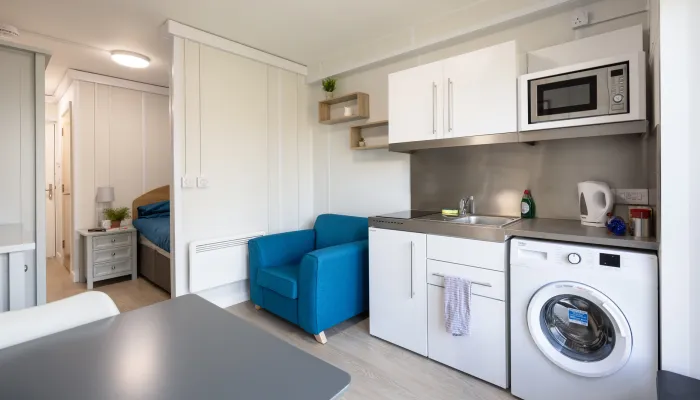 A compact studio room with a kitchenette, washing machine, microwave, sink, blue armchair, and a bed visible through an open doorway.