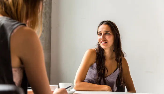Two people sit across from each other at a white table in a bright indoor setting. One person is in the foreground, partially out of focus, while the other is seated facing the camera. White coffee cups, a notebook, and a pen are on the table, suggesting a casual conversation or meeting. Natural light illuminates the scene against a plain light-colored wall.