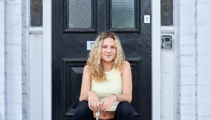 A young woman sits on the doorstep outside her home, holding house keys and looking peaceful