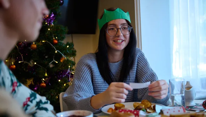 A young person reads a joke from a Christmas cracker over dinner, with a sparkling Christmas tree in the background