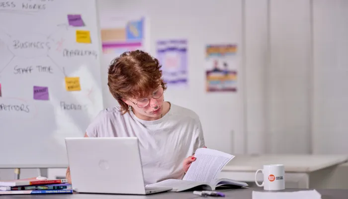 A young person studies with a book and laptop at their desk