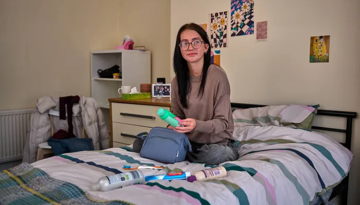 A young person sits on a bed holding personal hygiene items like shampoo